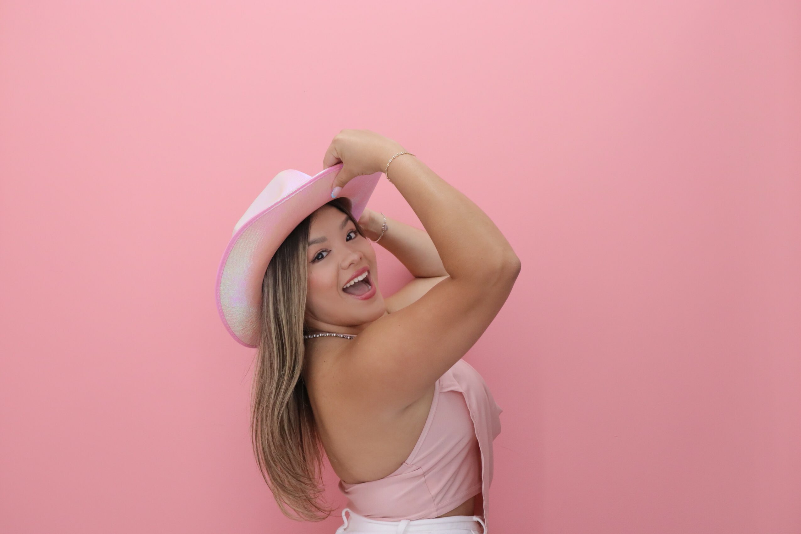 Woman wearing a pink dress and hat, smiling and posing with her arm raised against a pink background.