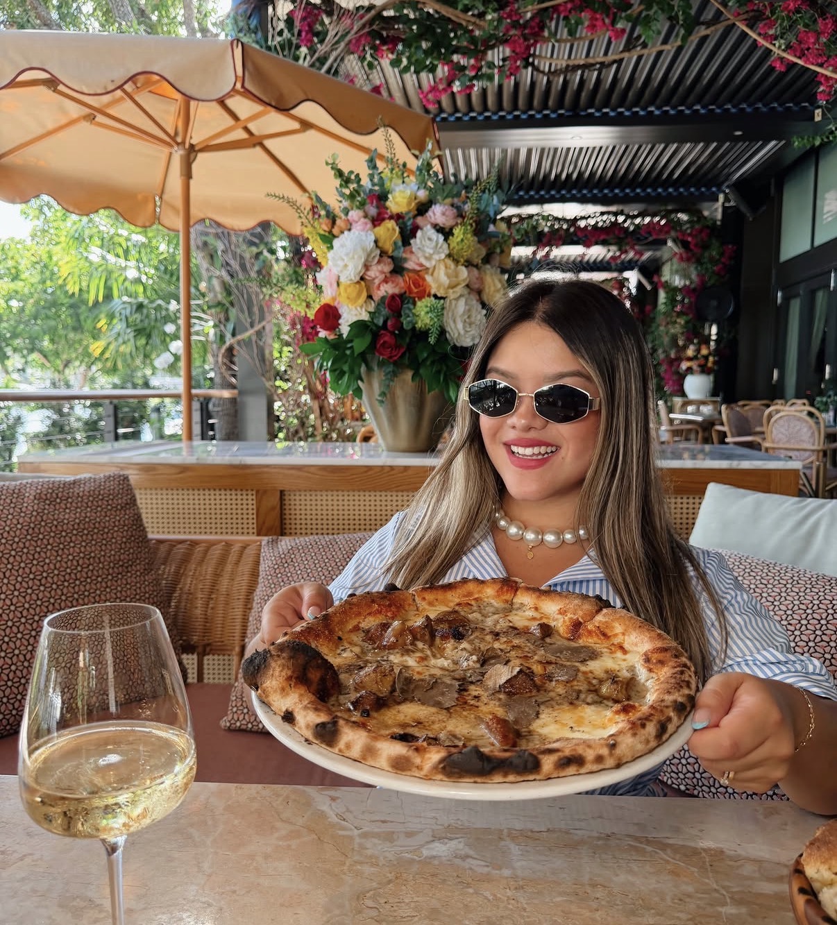 Woman with sunglasses smiling at a table holding a pizza, with a glass of white wine nearby, in a restaurant setting.