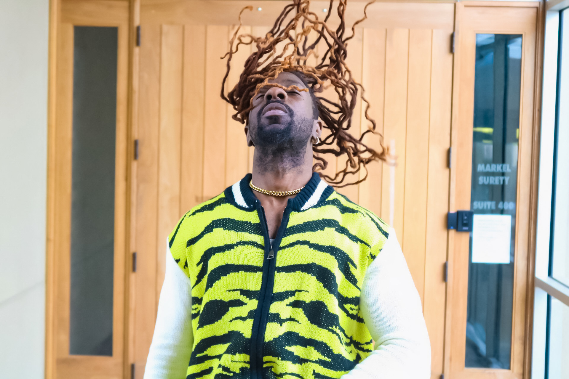 Person with long hair and glasses, wearing a yellow and black tiger-striped jacket, head tilted back, hair flying upward.