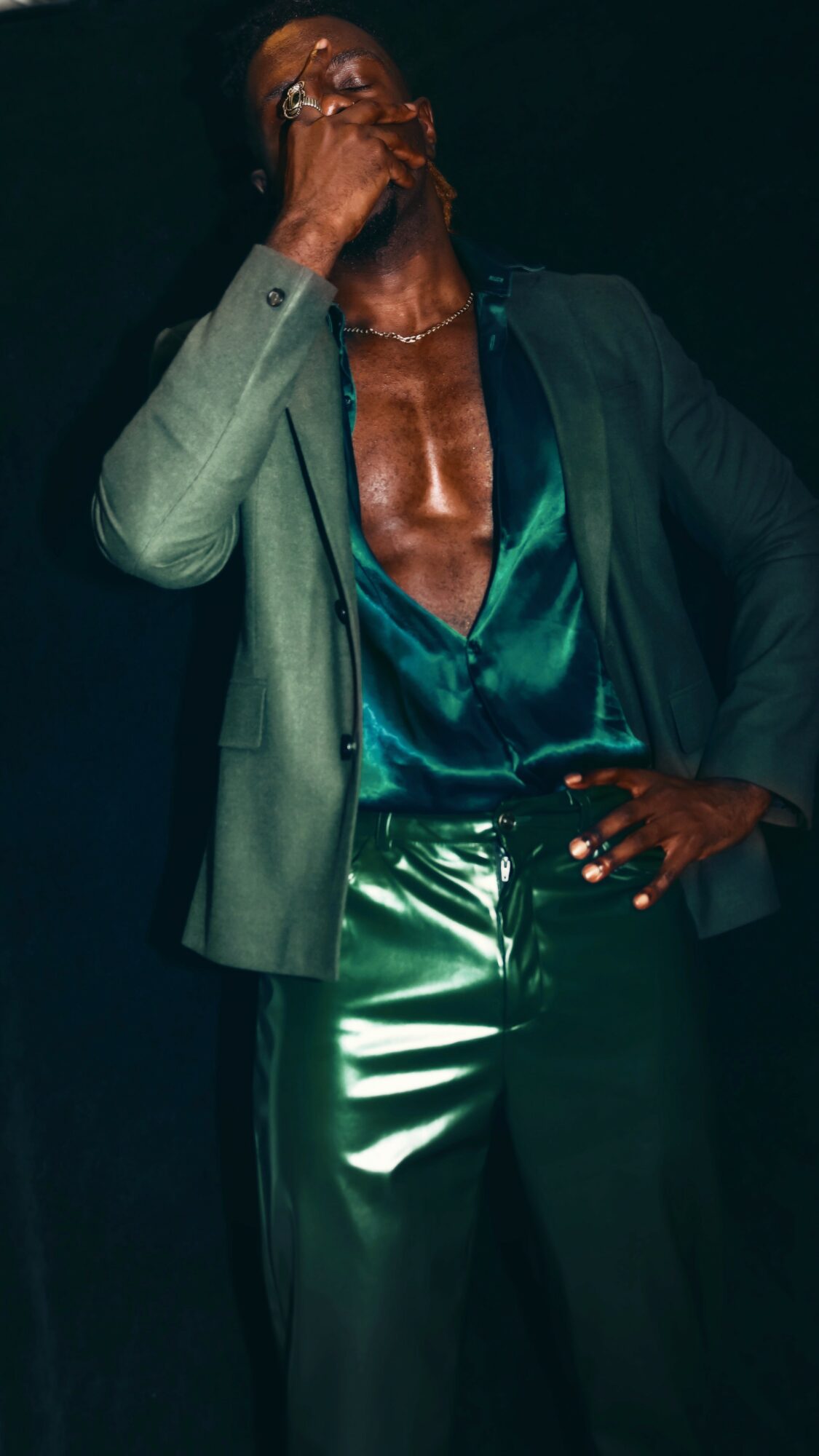 Person with hand covering mouth, wearing a blazer and shiny teal shirt, against a dark background.