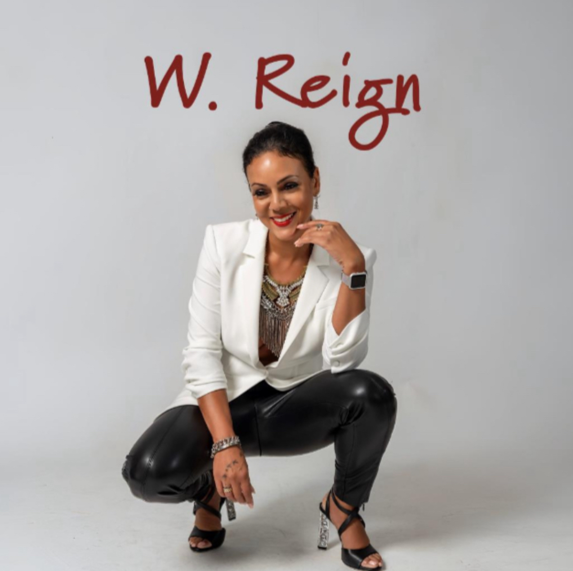 Woman crouching, smiling, wearing a white blazer, black pants, and high heels, with W. Reign written above her head.