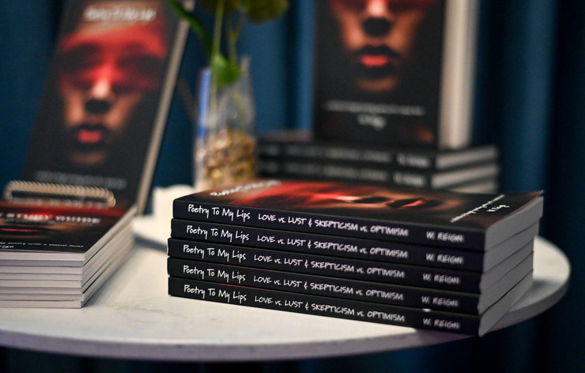 Stack of books titled 'Love & Lust' on a table with a blurred background of more books and a vase with flowers.
