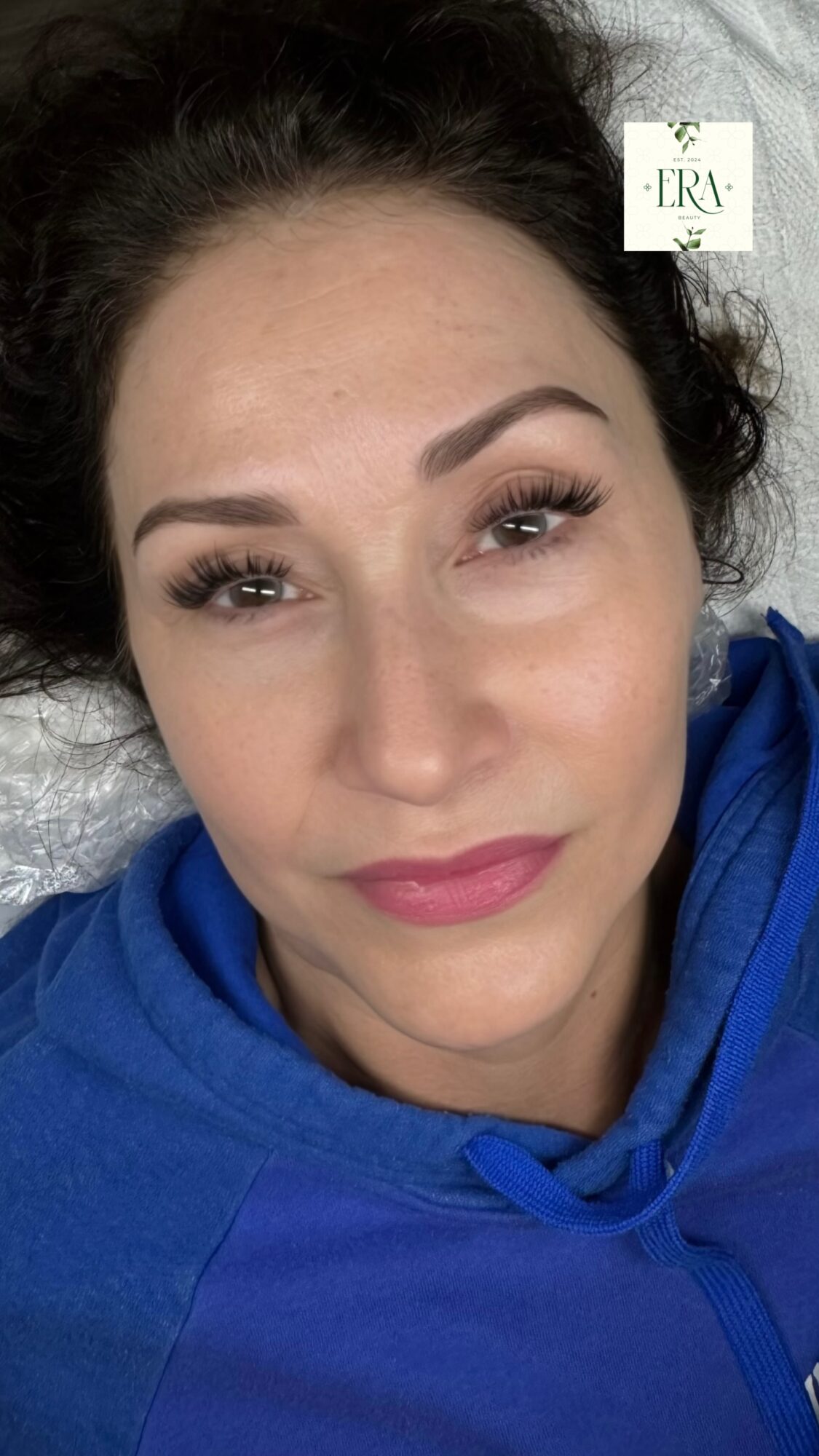 Close-up of a woman with dark hair, makeup, and a blue hoodie, lying down on a surface.