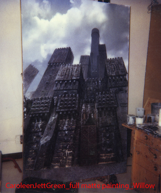 Painting of a tall, dark building with a cloudy sky background, leaning against a wall in a room. Wooden furniture and art supplies are nearby.