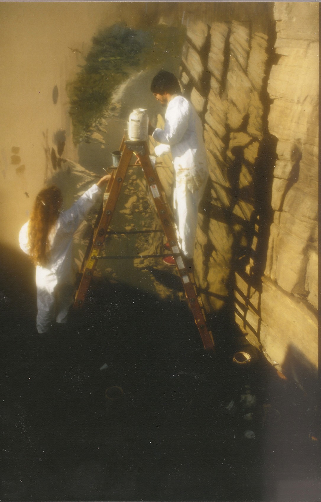 Two people in white clothing working on a ladder against a wall, with shadows cast on the ground.