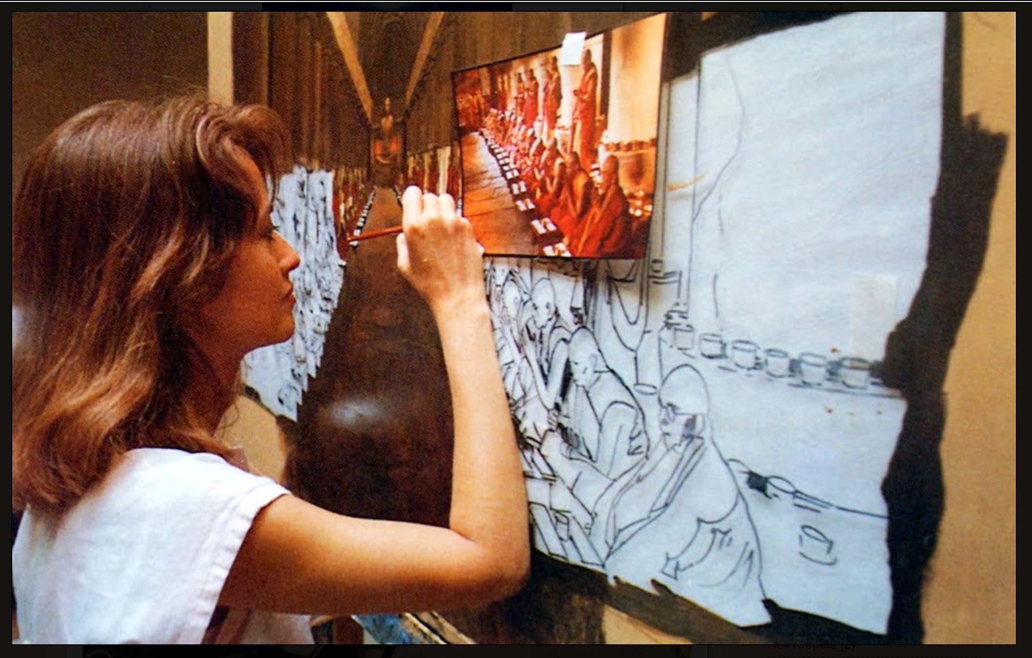 Woman painting a landscape on a canvas, with a reference photo above her, in an indoor setting.