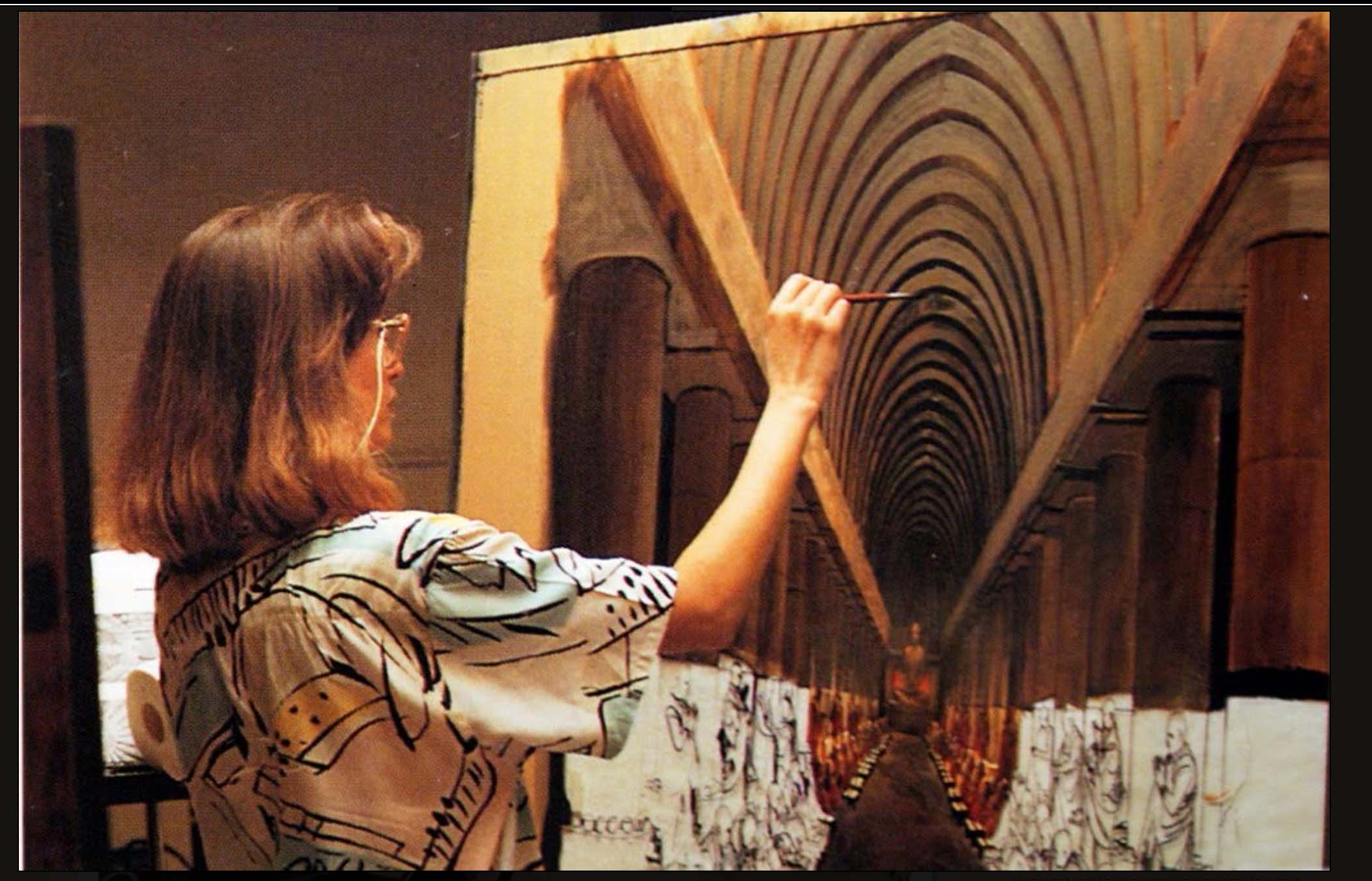 Woman with shoulder-length hair and glasses pointing at a large, curved, tunnel-like structure on a wall.
