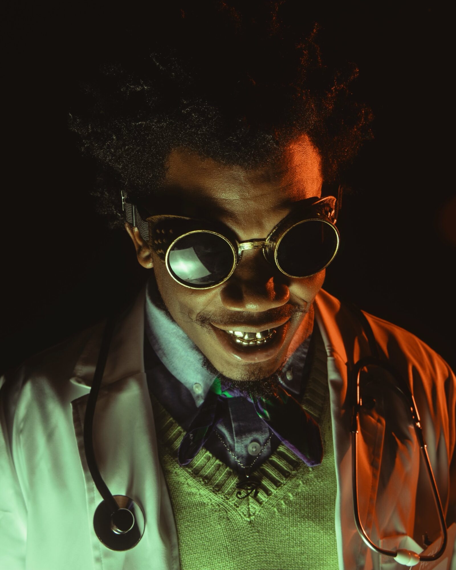 Person wearing round sunglasses, a white coat, and a stethoscope, smiling with visible teeth, in a dark setting.