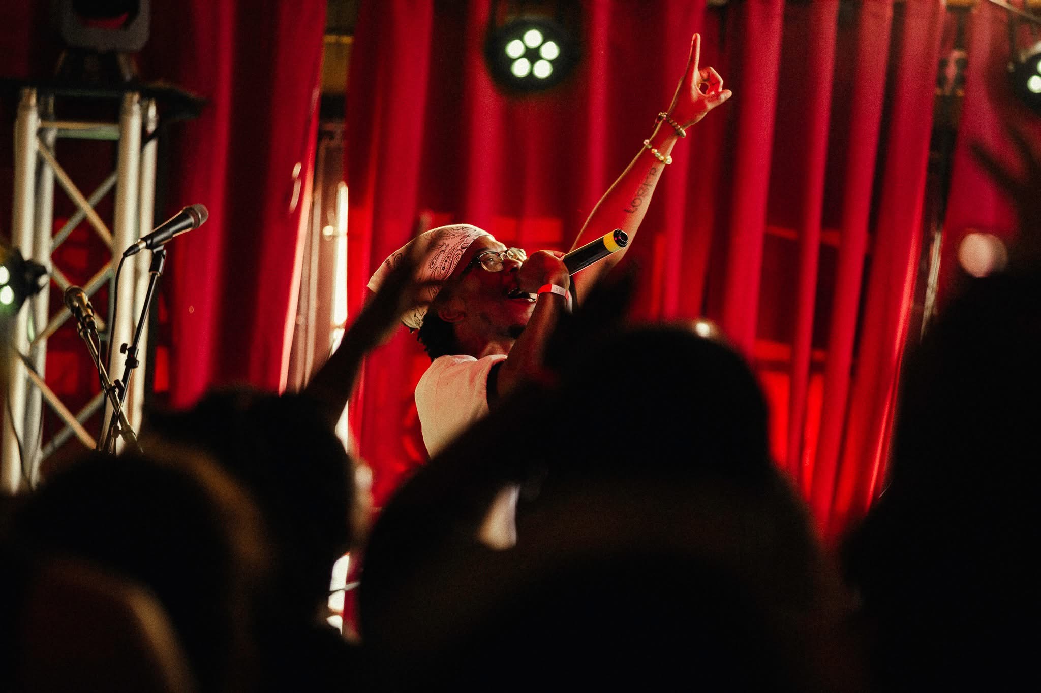 Performer on stage with red curtains, raising one arm, audience silhouettes in foreground.