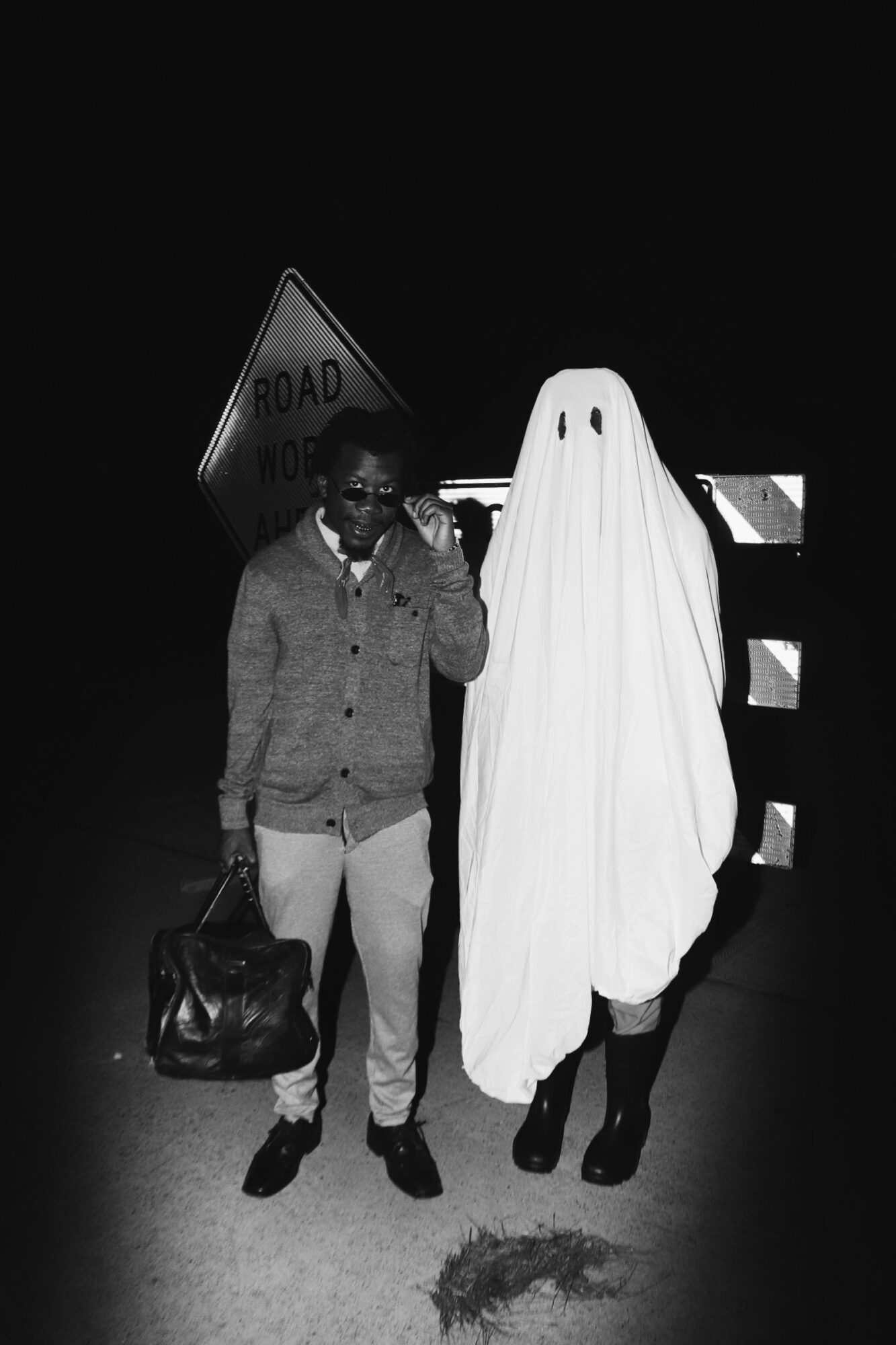Person walking with a bag next to someone dressed as a ghost on a dark road at night.