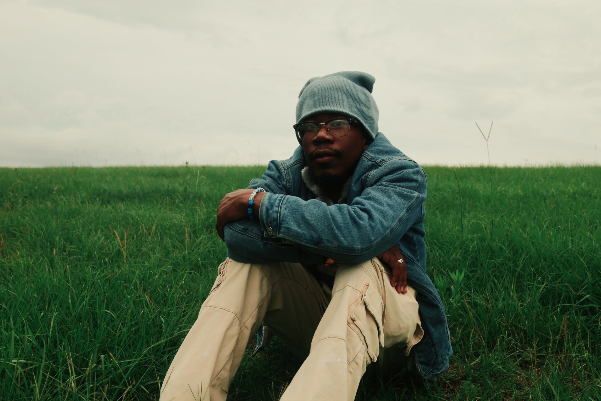 Person sitting on grass in an open field, wearing a beanie, glasses, denim jacket, and beige pants.
