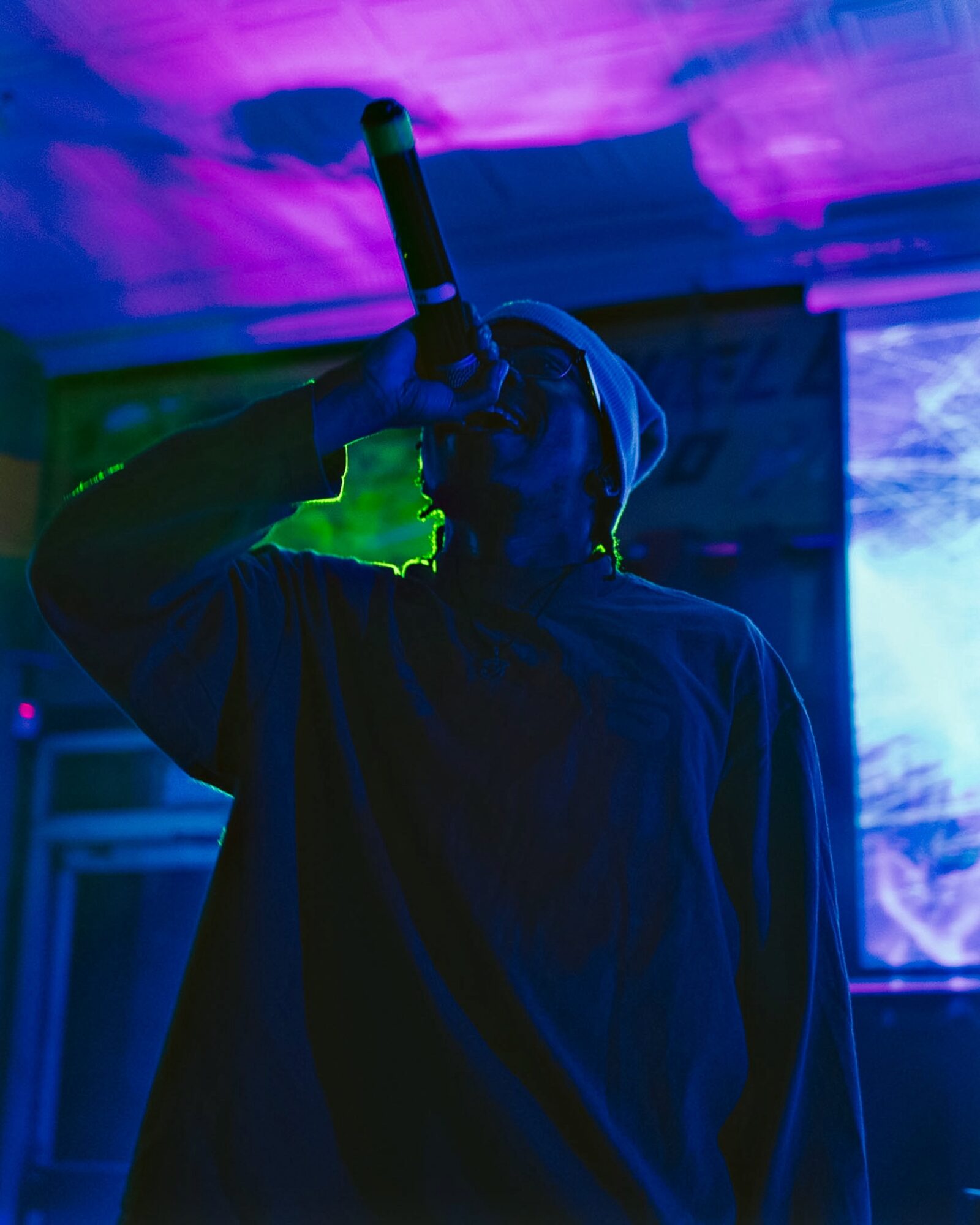 Person wearing a hoodie, holding a microphone, performing on stage with colorful lighting.