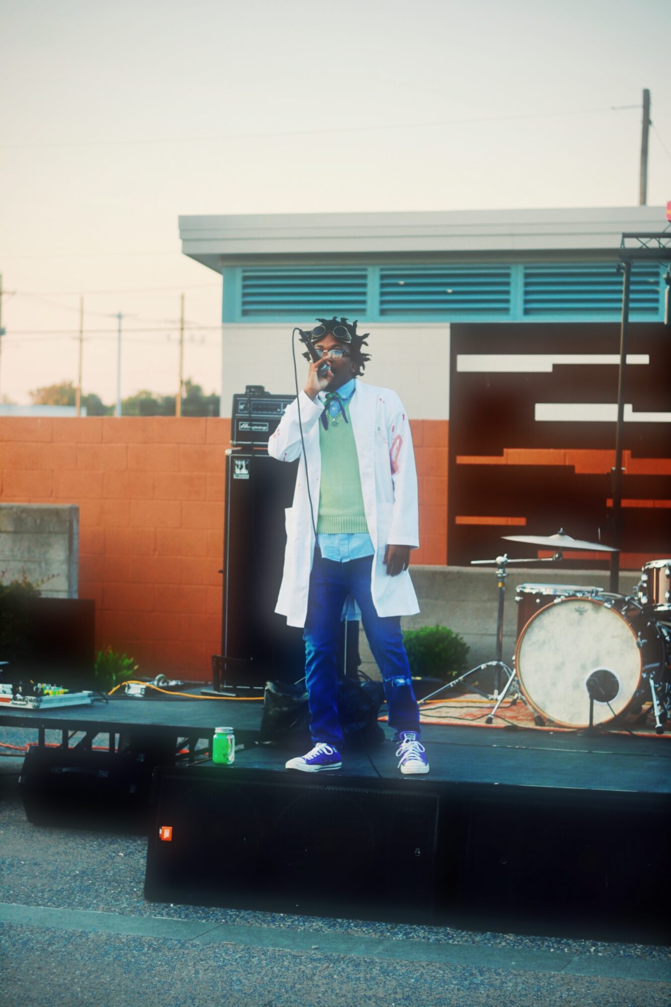 Person with dreadlocks and glasses standing on stage with microphone, wearing a white coat and colorful clothing, outdoors.