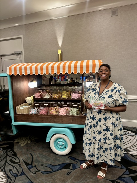Woman in floral dress standing next to a colorful food cart with striped canopy, smiling, holding a tray of food.