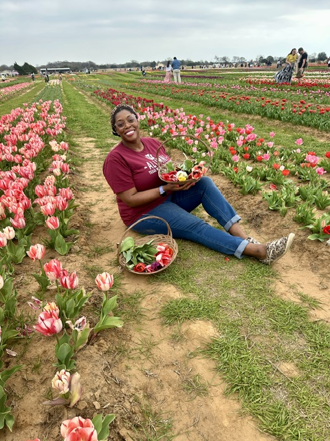 Woman sitting on a dirt path in a tulip field, smiling, holding a basket of tulips, with more flowers and visitors in background.