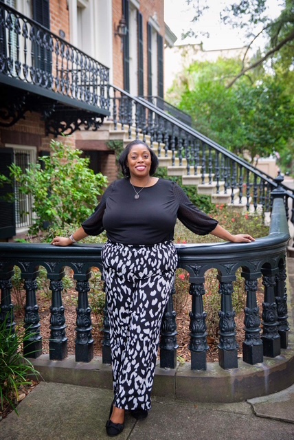 Woman standing outdoors on a curved black railing, with greenery and a building in the background.