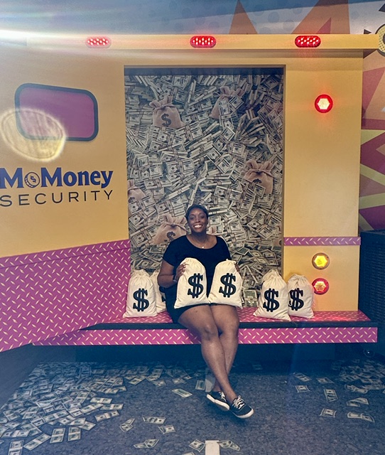 Person sitting on a pink bench surrounded by money bags, with a money-themed backdrop and colorful decorations.