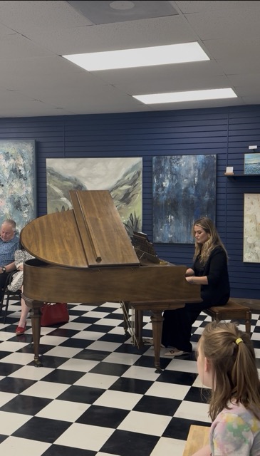 Woman playing grand piano in an art gallery with checkered floor and paintings on blue walls.