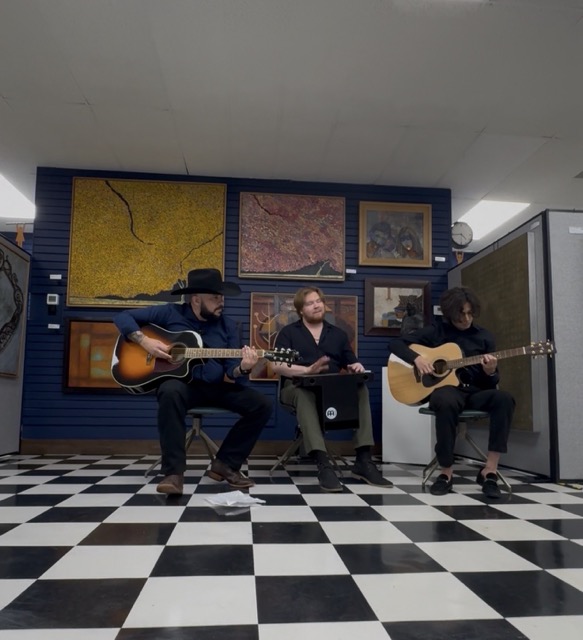 Three musicians playing guitars and a keyboard sit on chairs in a room with artwork on the wall and a checkered floor.