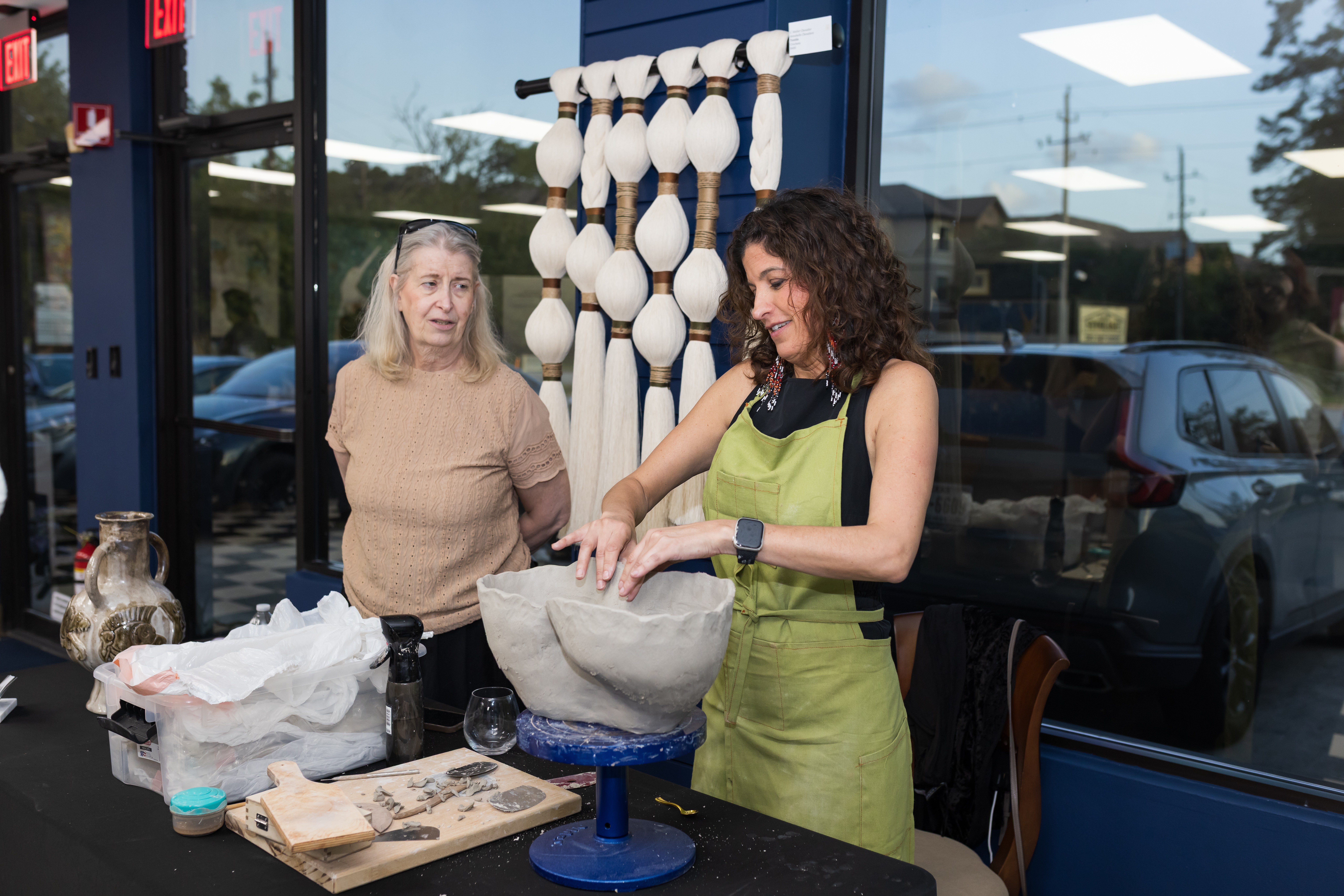 Two women working with a large stone bowl inside a shop, with decorative objects and a window showing parked cars outside.