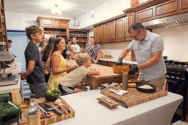Group of children and adults gathered around a kitchen counter watching a man prepare food, with wooden cabinets and kitchen appliances visible.