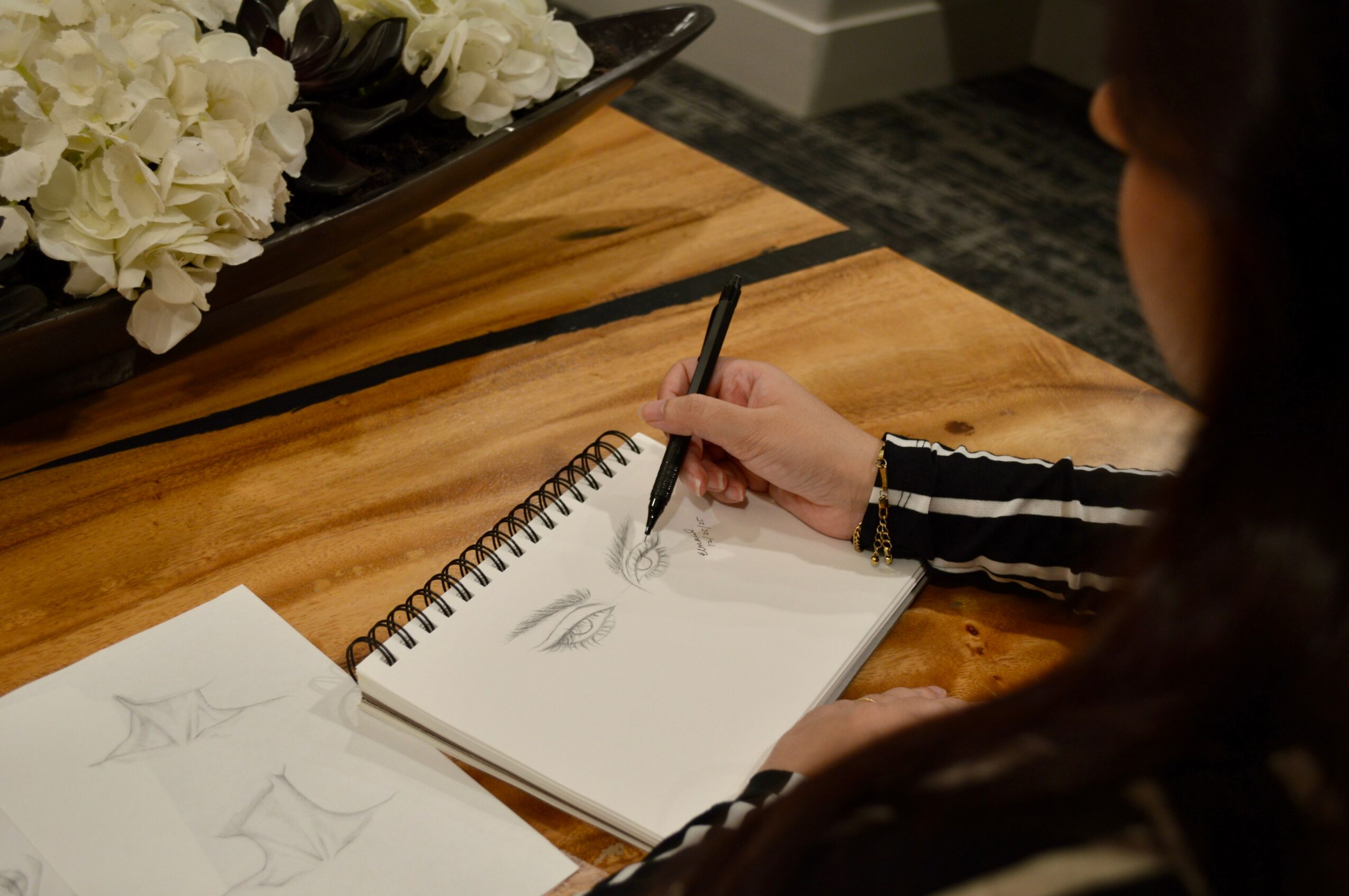Person drawing sketches in a spiral notebook on a wooden table, with flowers nearby.