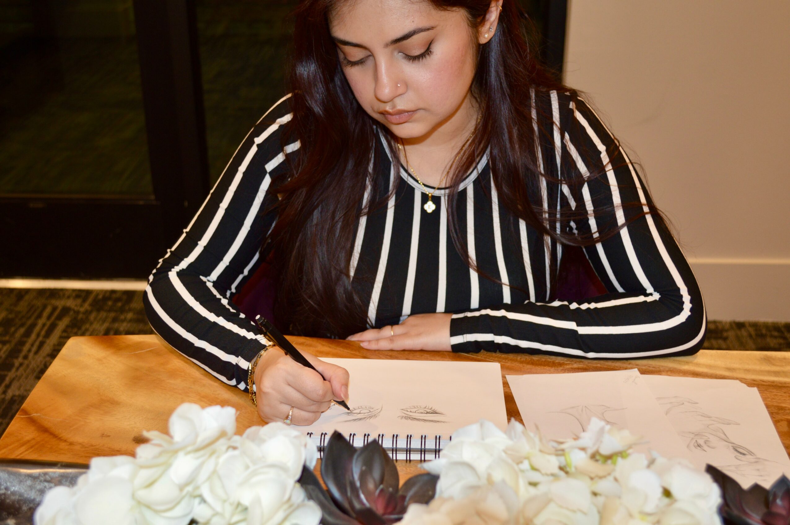 Young woman with long dark hair writing at a table with white flowers, papers, and a pen, indoors.