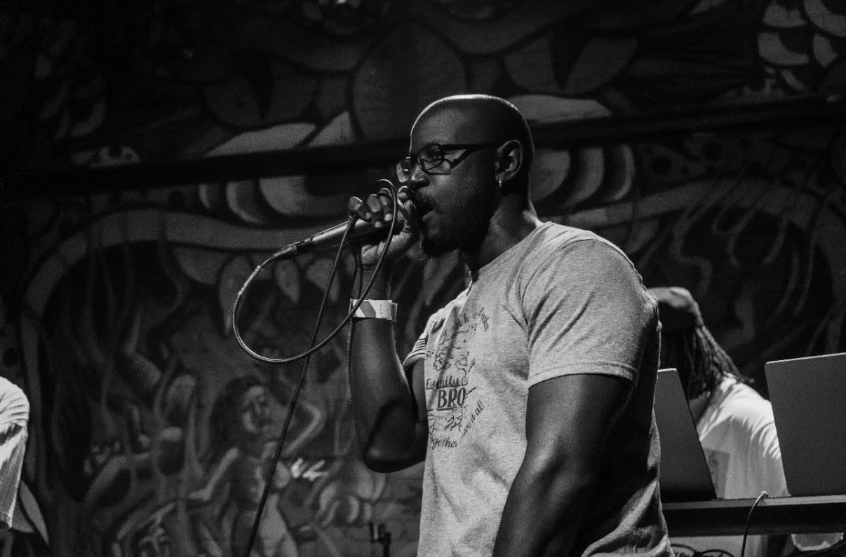A man with glasses and a beard sings into a microphone on stage, with a mural background.