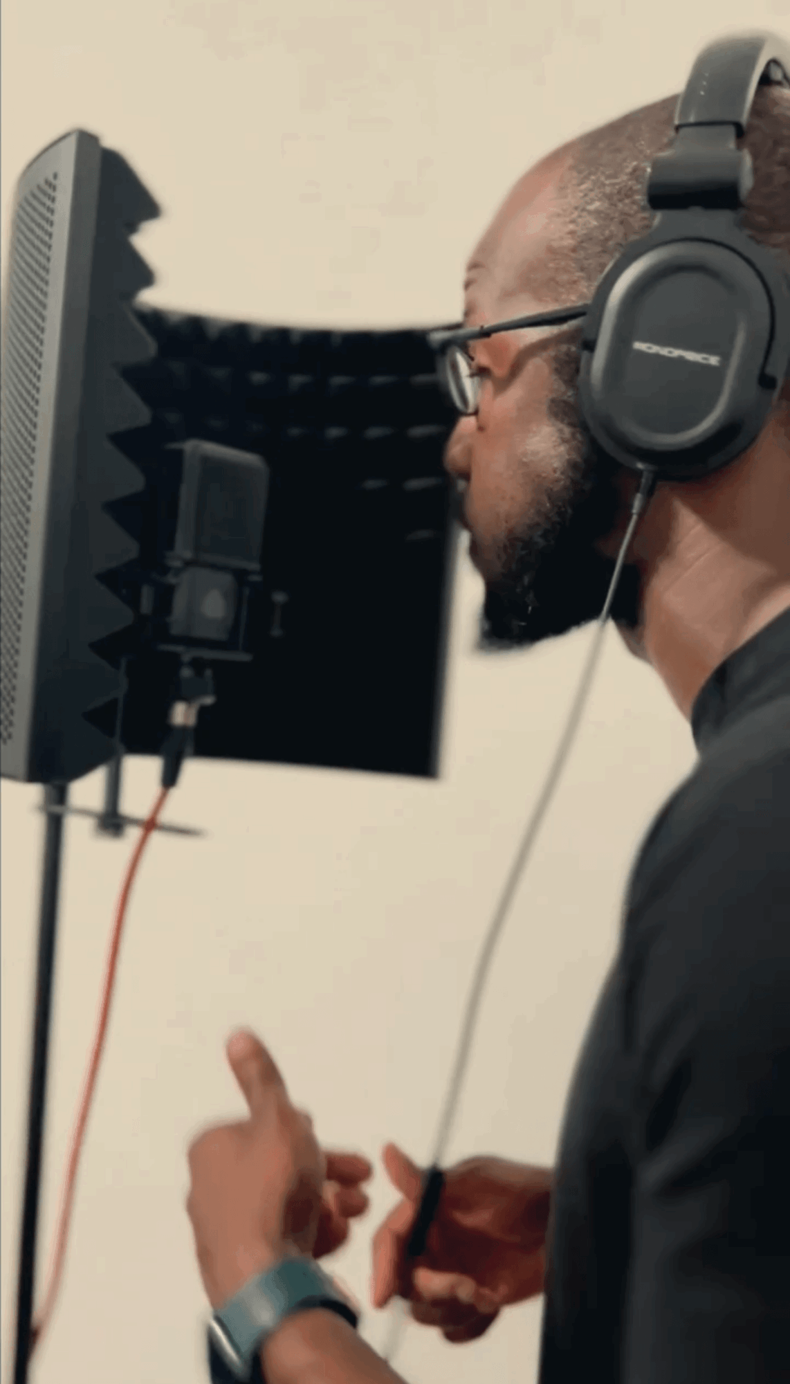 Person recording vocals in a studio with headphones, microphone, and soundproofing panel, facing right side, with hand raised.