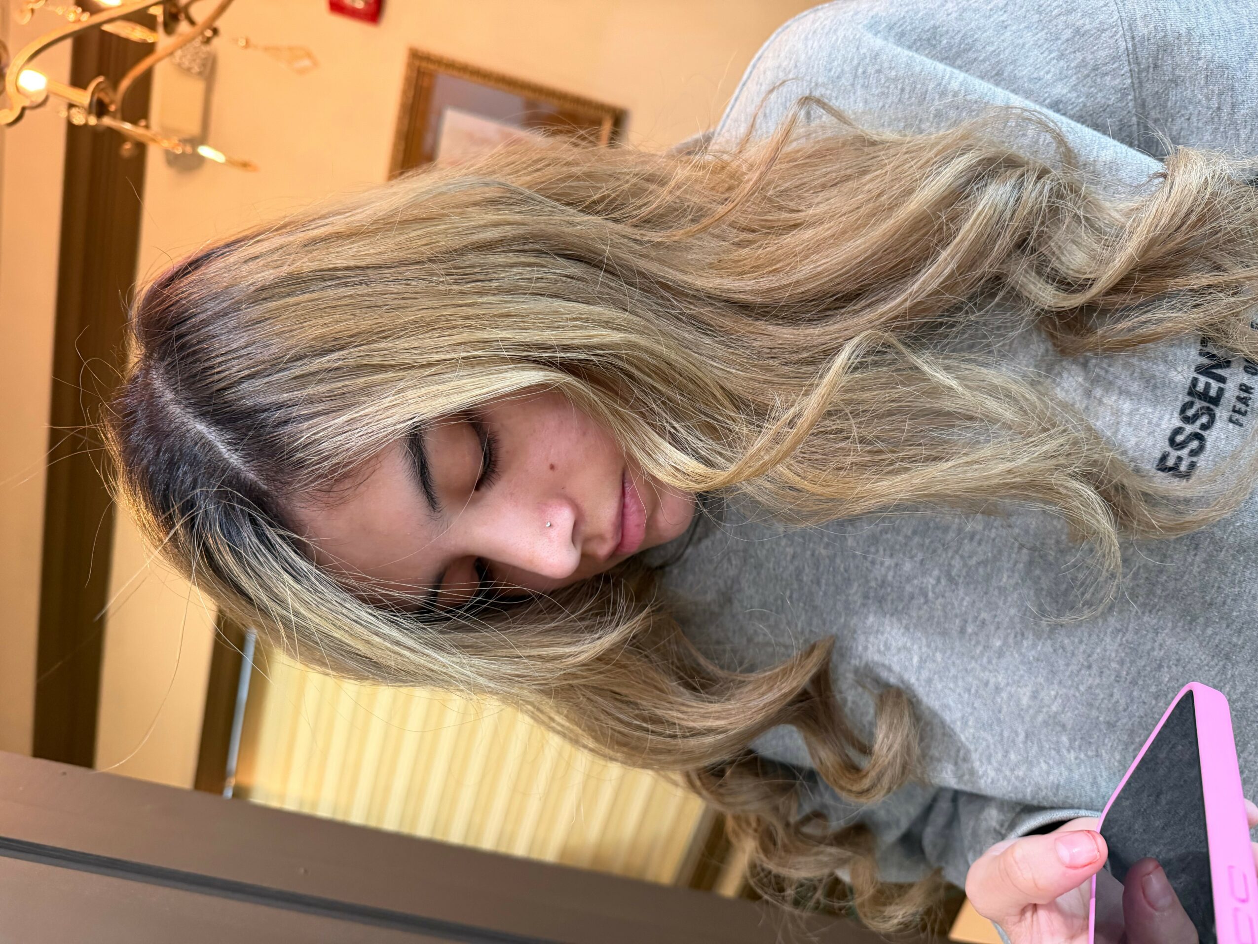 Young woman with long, wavy blonde hair looking down, holding a pink phone, indoors with warm lighting.