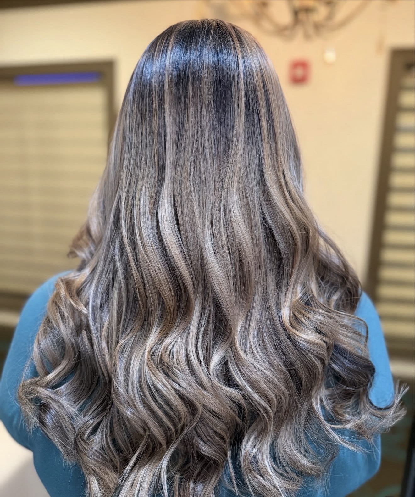 Back view of a person with long, wavy, multi-toned hair in shades of brown and blonde, wearing a blue top.