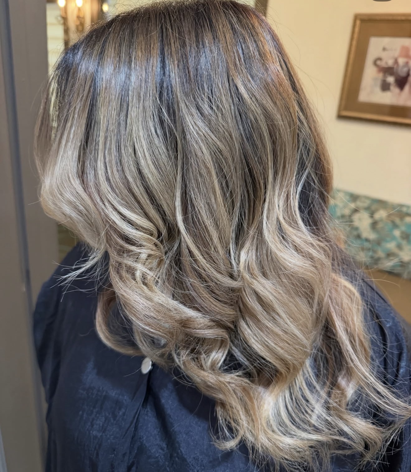 Wavy, blonde hair with darker roots, styled in loose curls, seen from the back in an indoor setting.