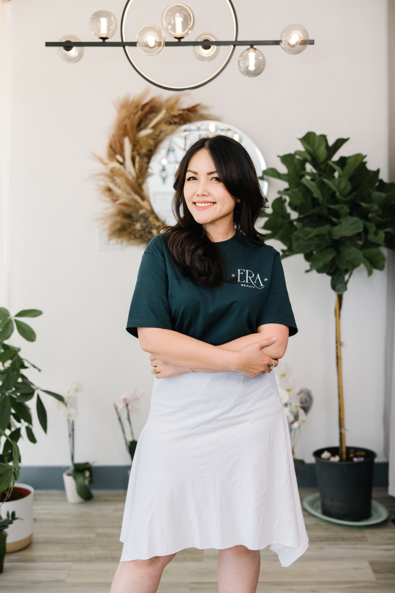 Woman with dark hair smiling, standing indoors with plants and a mirror behind her.
