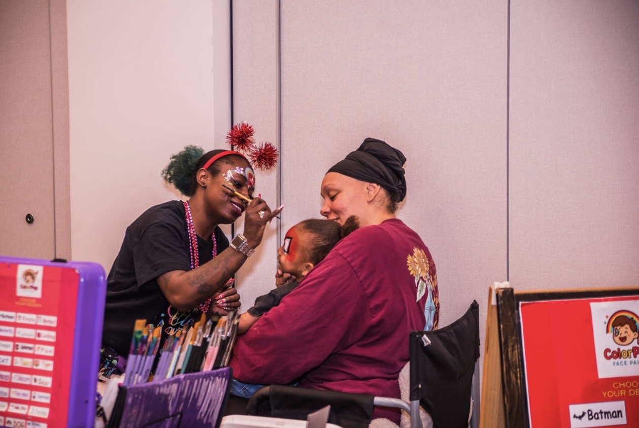 Two women and a child sit together, smiling and engaging in a joyful moment indoors, with colorful books and posters nearby.
