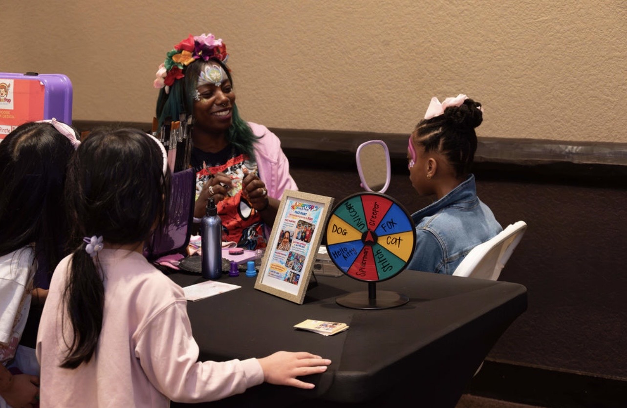 Group of children and a woman with colorful hair and flower crown sitting at a table with a spinning wheel and picture frame.