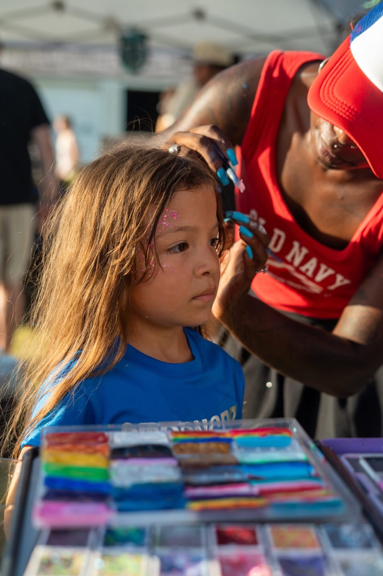 Child with long hair looking at colorful items on table, person in red cap and shirt nearby, outdoor setting