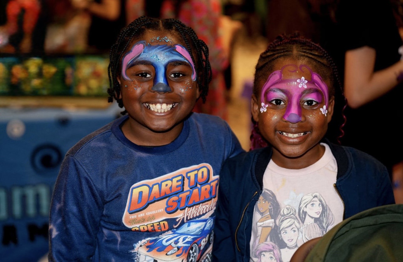 Two children with face paint smiling at a festive event, colorful background, wearing casual clothes.