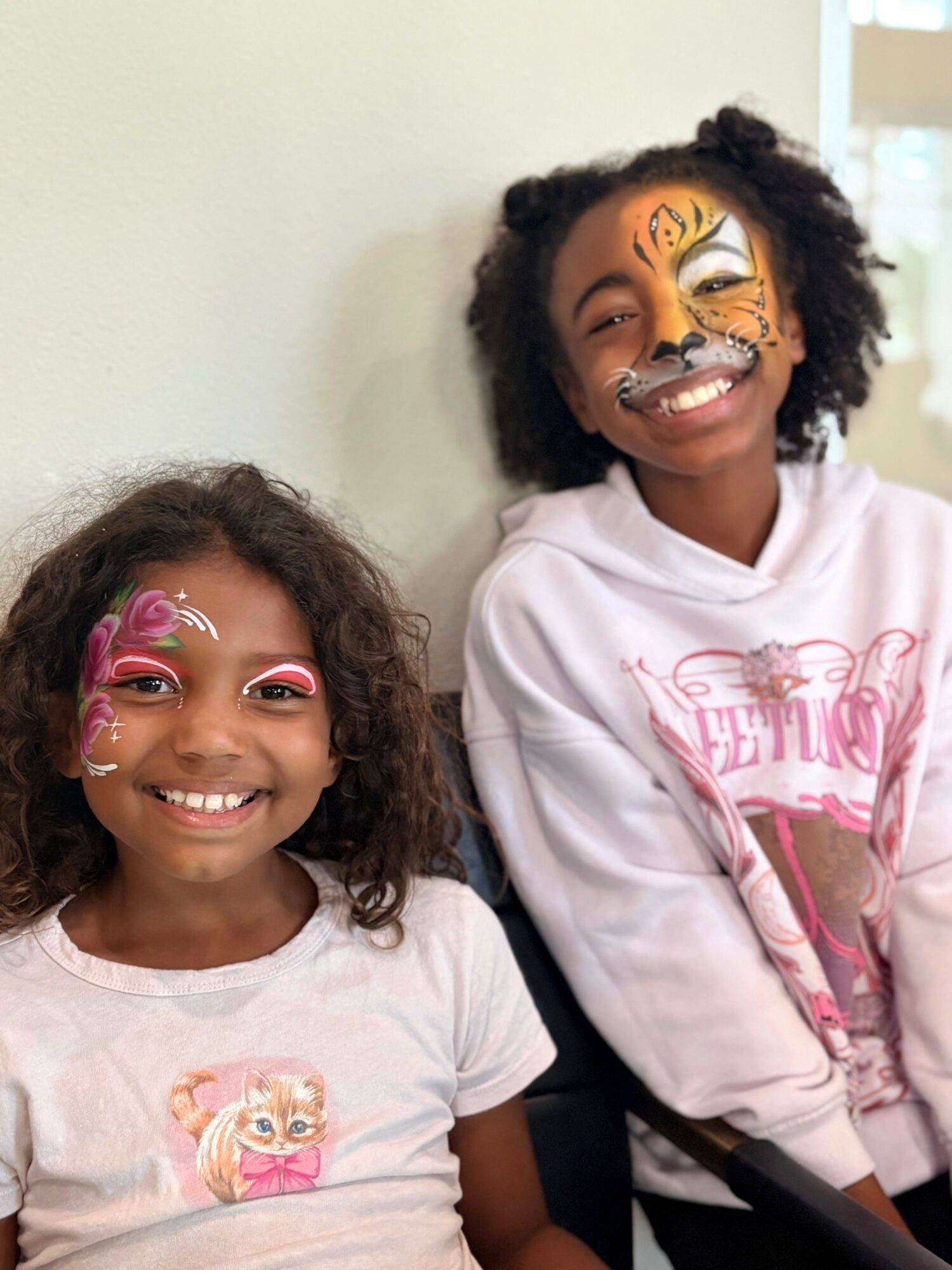 Two smiling children with face paint, one with tiger design and the other with butterfly, sitting against a plain wall.