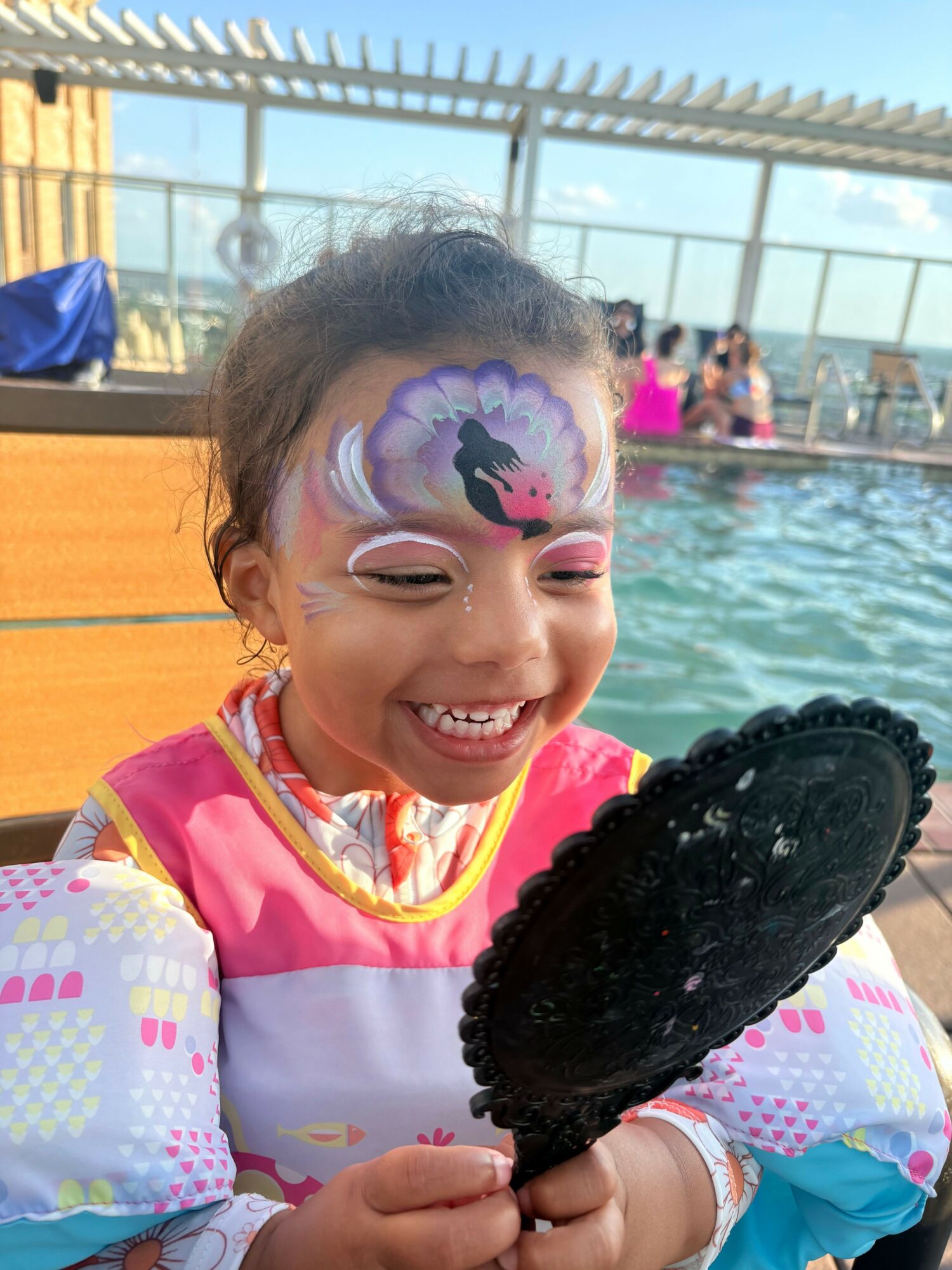 Child with face paint smiling, holding a black lace fan, at a poolside with people in the background.