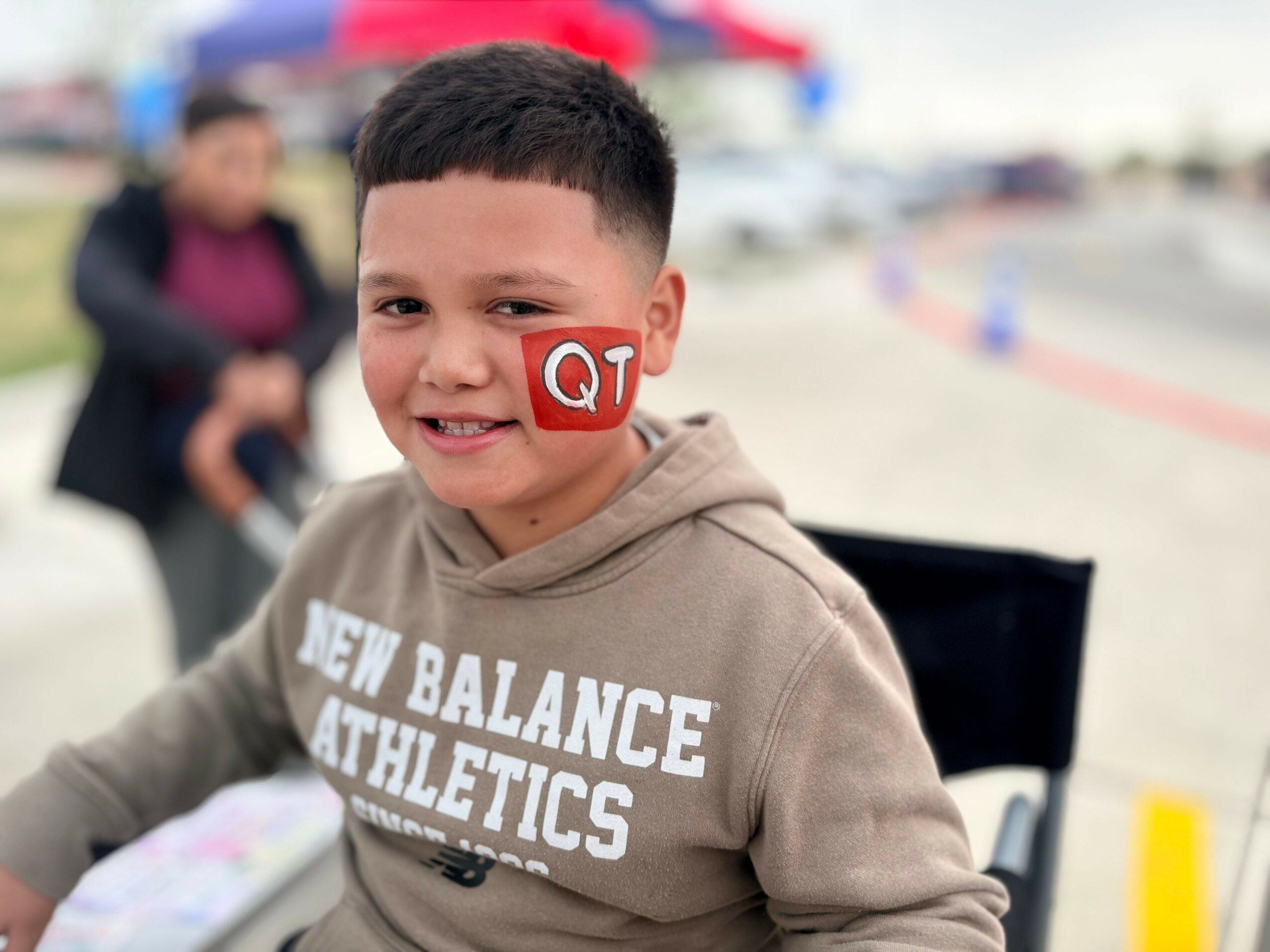Young boy smiling outdoors, wearing a beige hoodie with white text, with a blurred background of people and cars.