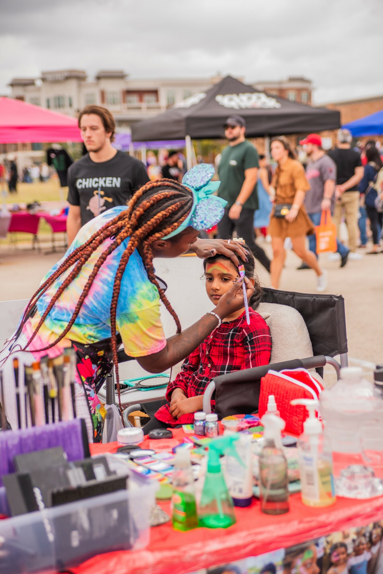 Child sitting at a table with colorful items, a woman with braided hair applying face paint, outdoor market scene with people and tents
