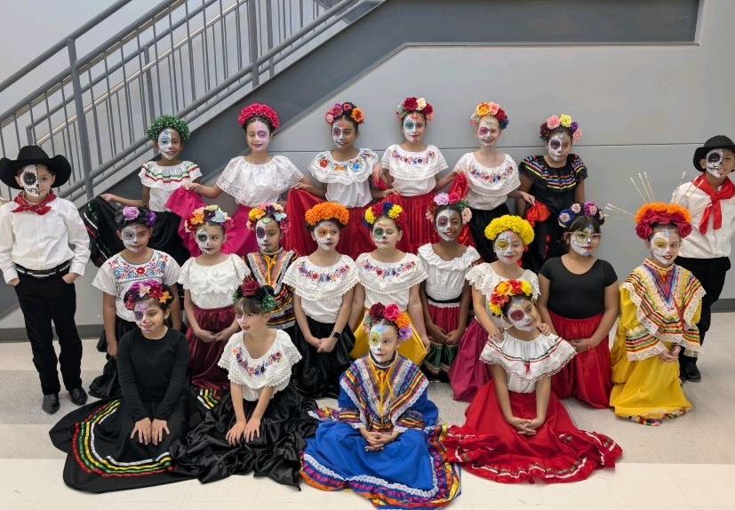 Group of people in colorful traditional costumes, some with floral headpieces, posing indoors against a staircase.