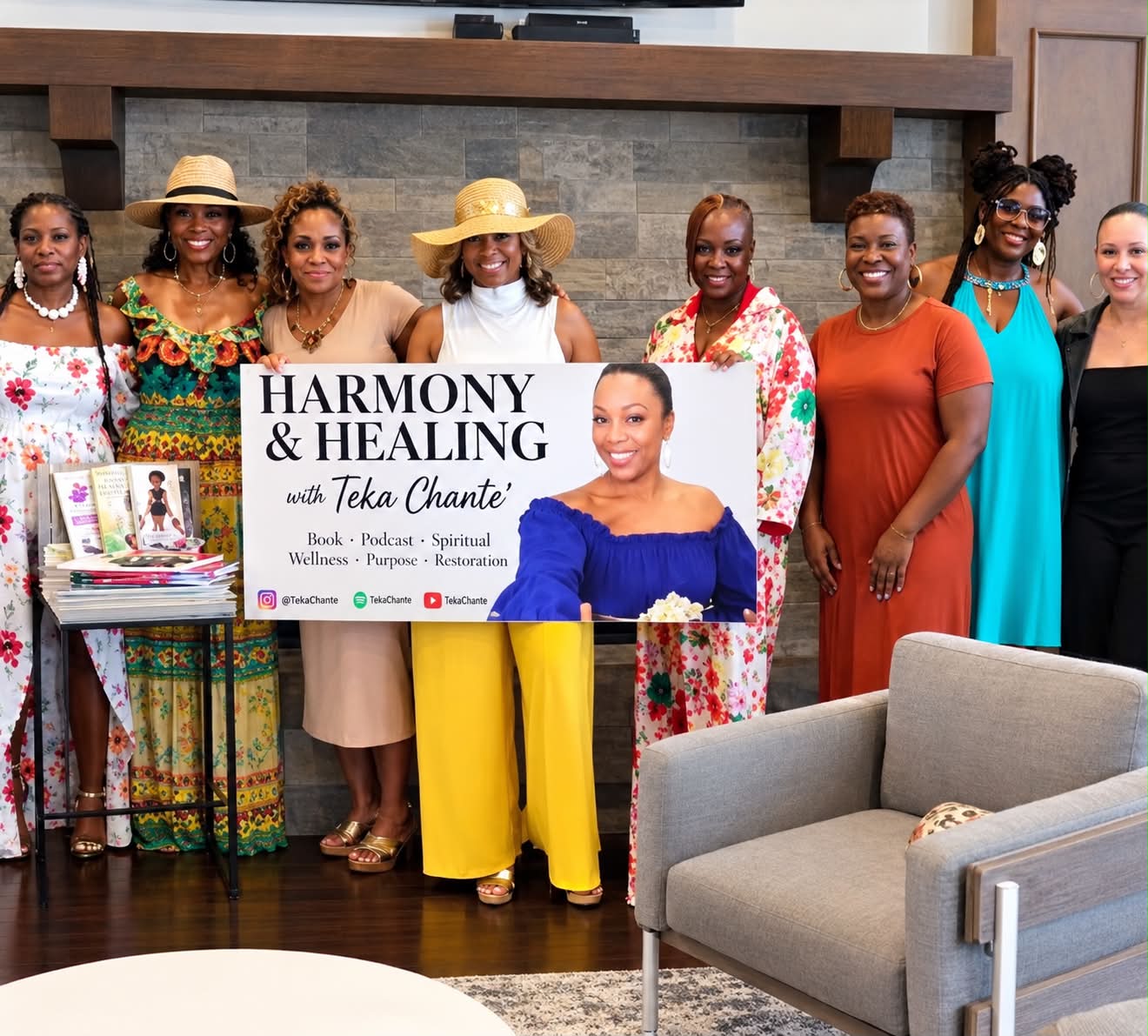 Group of women standing indoors, holding a sign that reads 'Harmony & Healing with Teka Chante,' with a fireplace in the background.