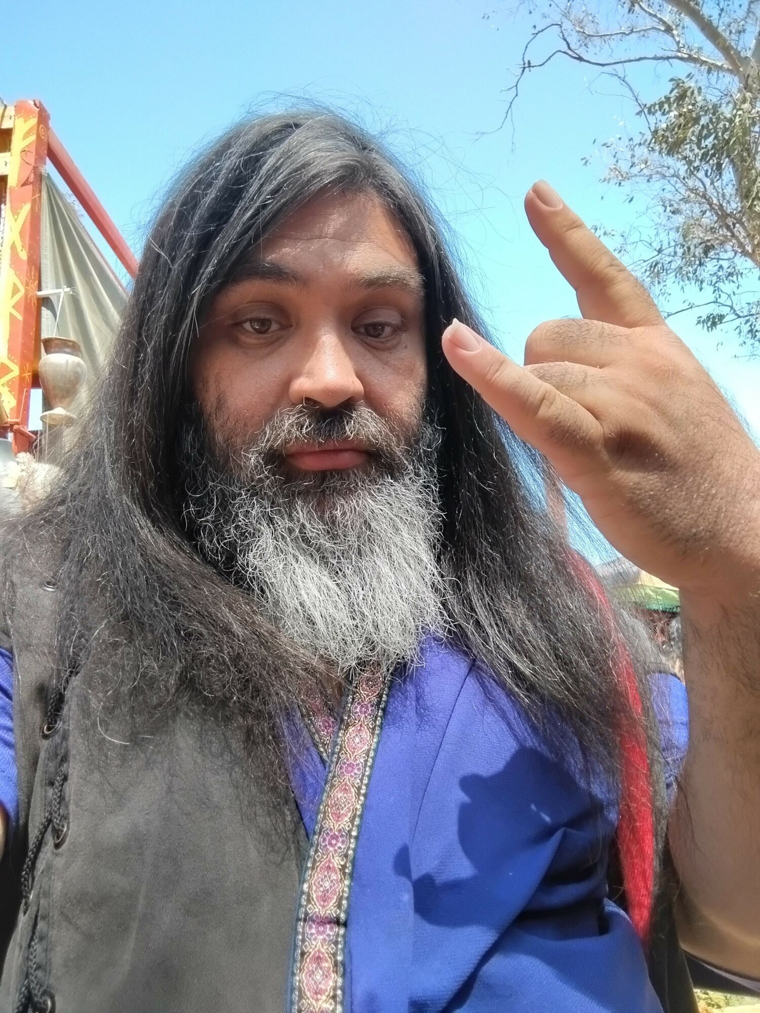 Man with long gray hair and beard making a hand gesture outdoors, blue sky and tree in background.