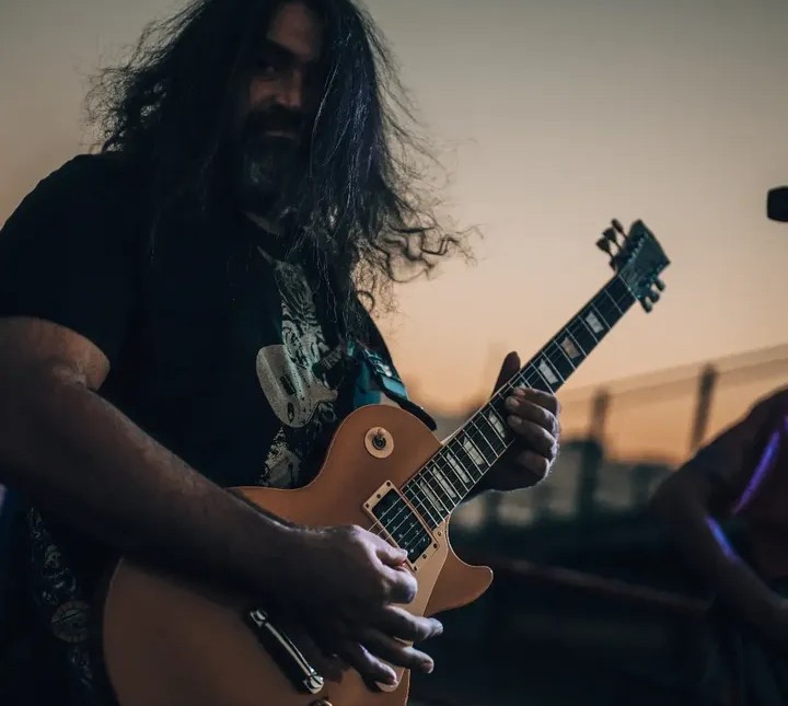Person with long hair playing an electric guitar outdoors during sunset, smiling, with a blurred background.