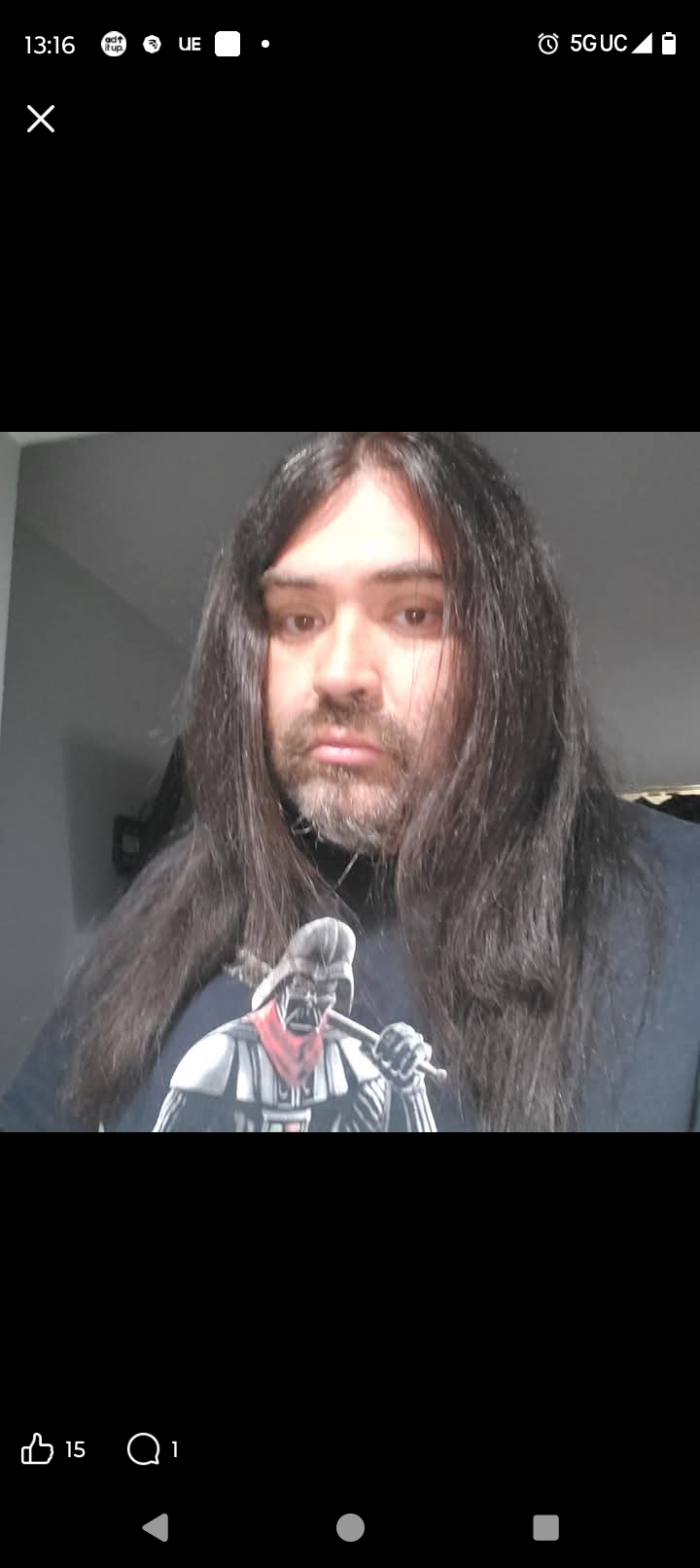 Person with long dark hair and beard wearing a graphic t-shirt, looking at the camera indoors.