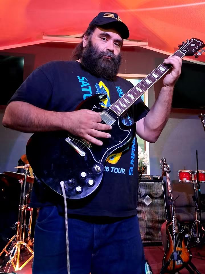 Man with a beard and cap playing an electric guitar on stage, with musical instruments in the background.