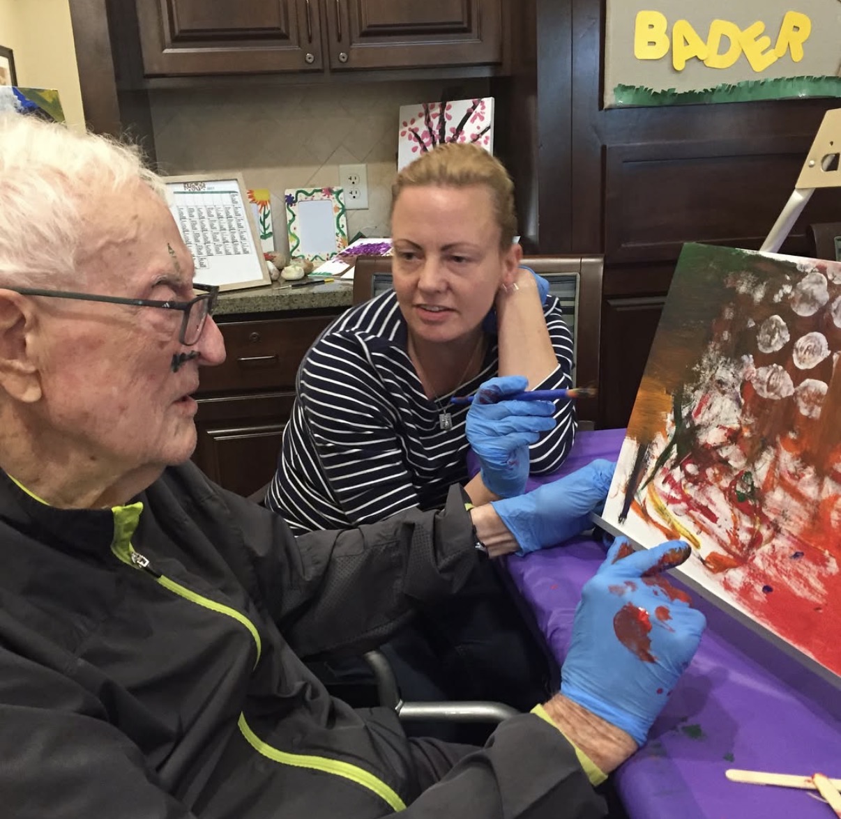 An elderly man and a woman with gloves paint on a canvas in a room with cabinets and colorful decorations.