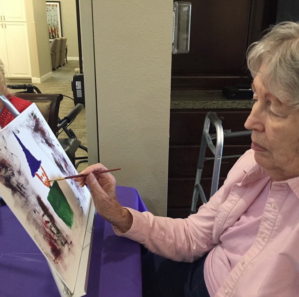 Elderly woman painting on canvas with a brush, seated at a table, wearing a pink shirt, in an indoor setting.