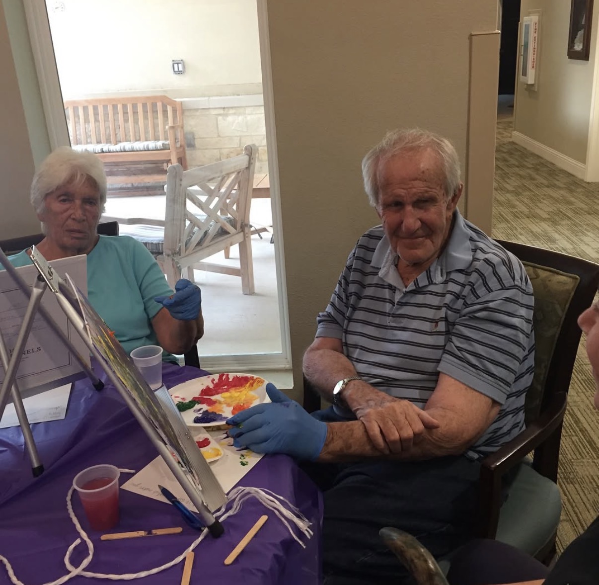 Two elderly people sit at a table with art supplies, one smiling, in a room with a window and outdoor view.
